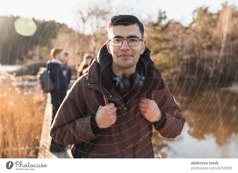 Smiling ethnic man looking at camera near lake in autumn forest smile nature park hike traveler friend positive together friendship content glad male young