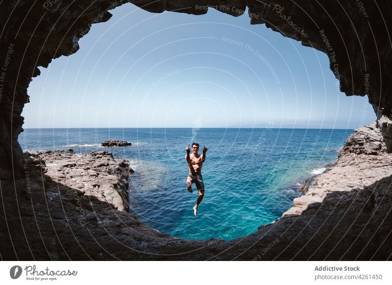 Smiling man showing fuck gesture while jumping over sea ocean cave content nature highland hole sky activity energy smile rude middle finger rebel aggressive