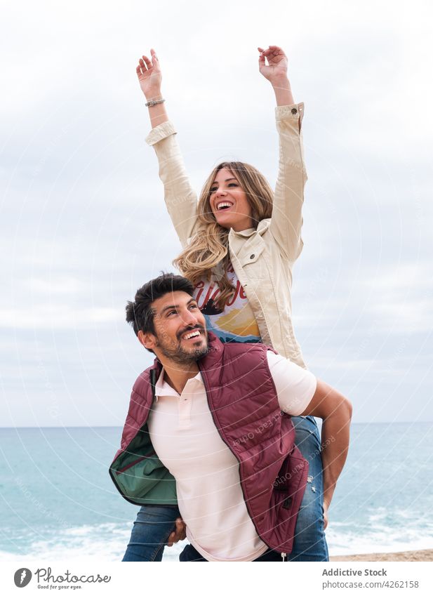 Happy ethnic man giving girlfriend piggyback ride against sea boyfriend relationship happy love romance arms raised portrait sky nature ocean smile shore couple