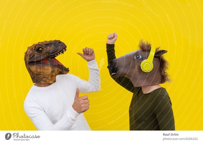 Funny couple of happy young man and woman with dinosaur and horse head mask dancing. Positive and euphoric people lifestyle, isolated on yellow background in studio with blank wall for copy space .