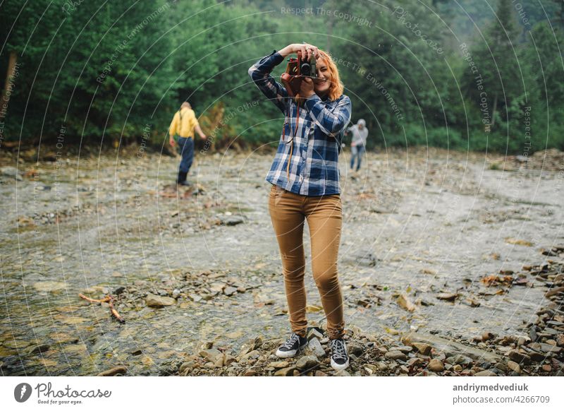 Young redhead woman with vintage camera takes photo near the river in a mountains travel retro young beautiful summer nature female photographer girl outdoor