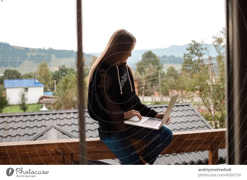 Closeup image of a woman working and typing on laptop while sitting on wooden balcony with green mountains on foggy day with blue sky background closeup travel