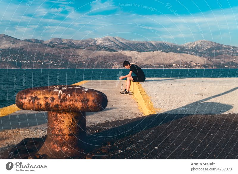 pensive teenager sitting on a pier by the sea with beautiful scenery in the background young man sad serious alone lonely bottle water harbor worried landscape