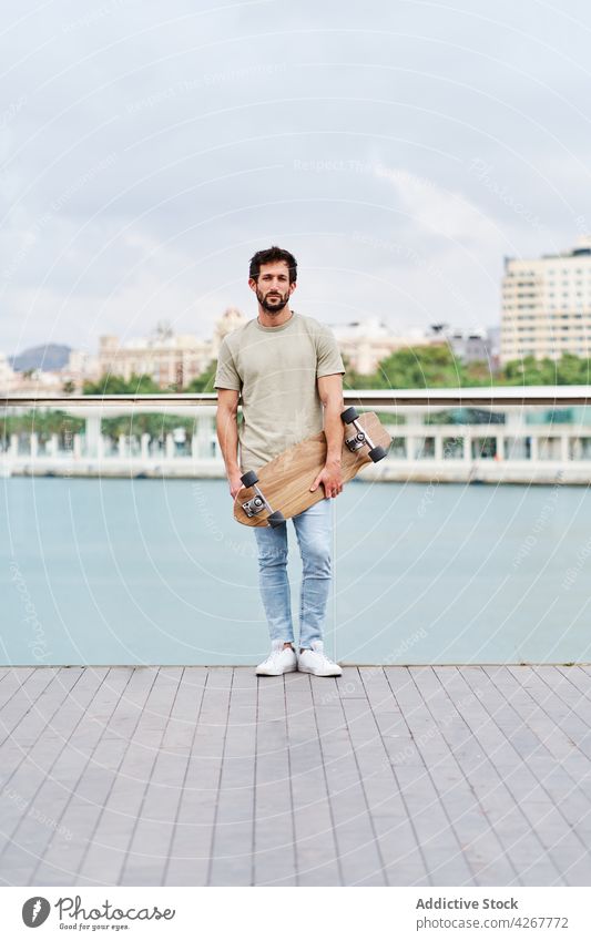 Bearded man with skateboard standing on city lakeside skater unemotional casual waterfront promenade embankment young river pond summer sunny town boardwalk