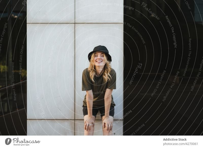 Candid woman touching knees against building wall candid smile touch knee lean forward charming friendly pleasant portrait cheerful enjoy casual wear content