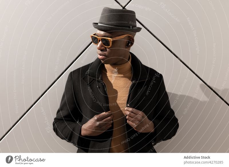 Stylish black man standing against white wall style outfit confident appearance trendy cool well dressed hat sunglasses individuality personality modern garment