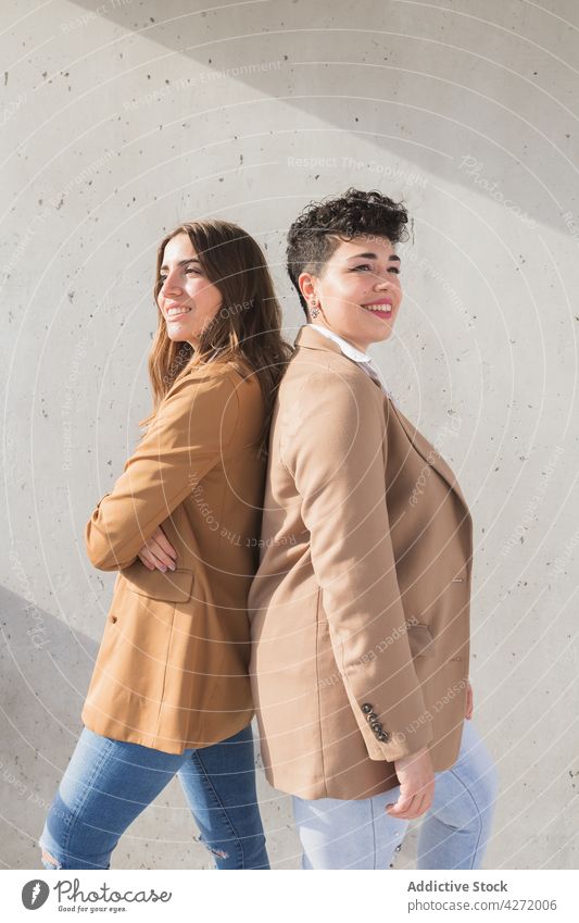 Happy girlfriends standing back to back near wall women smile street trendy confident sunny happy female young together friendship jacket jeans positive elegant