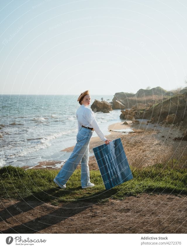 Young woman walking on seashore with painting in hand nature painter beach style coast female young trendy hat ocean holiday sand casual vacation outfit fashion