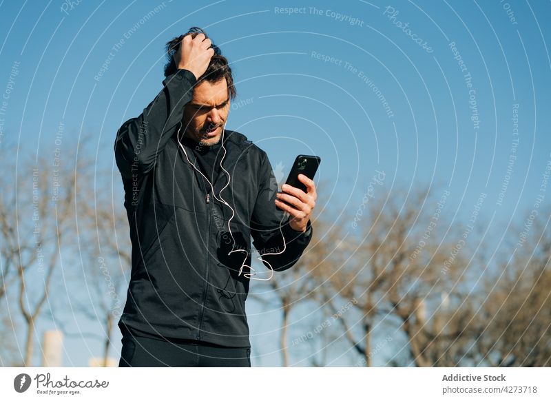 Sportsman in earphones browsing smartphone in city park sportsman chatting attentive listen music masculine using gadget device focus sportswear meadow natural