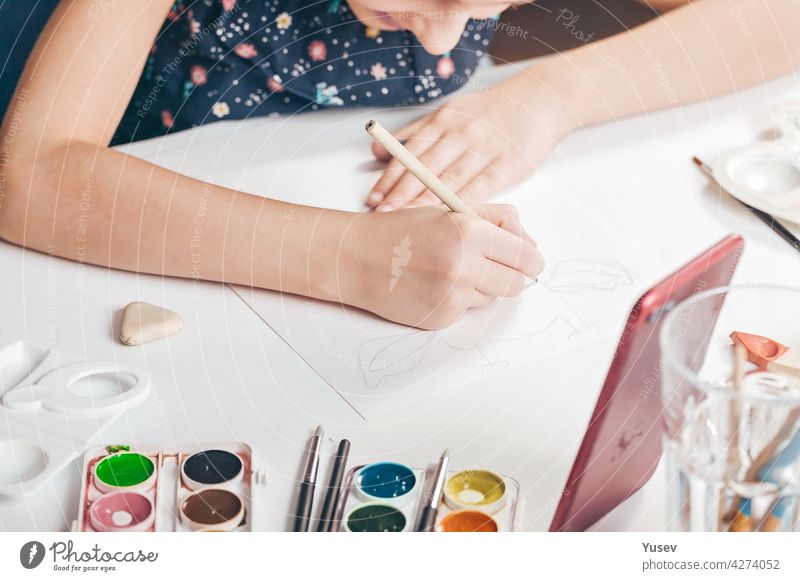 The hands of a cute caucasian girl are drawing a pencil sketch. A girl is watching an online lesson on a smartphone. Classes with children at home. Hands in the frame