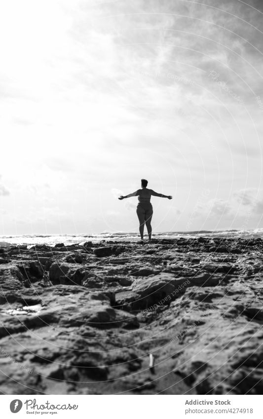 Anonymous woman raising hands on rocky shore near ocean sea arms raised plus size alone bw female plump sunny black and white cloud summertime wavy aqua tourist