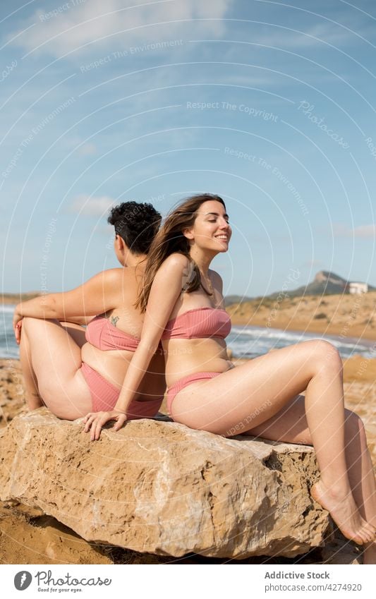 Women in swimsuits on rocky boulder on beach near ocean women sand sea joy blue sky friend swimwear female young summer vacation stone shore cloudy enjoy water