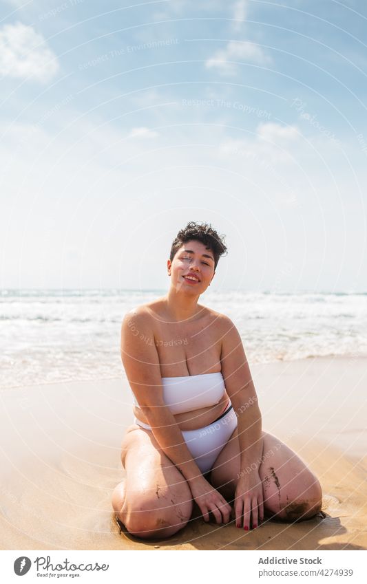 Lady sitting on sandy coast near sea woman beach ocean smile plus size swimsuit vacation blue sky happy female young aqua enjoy alone bright toothy smile