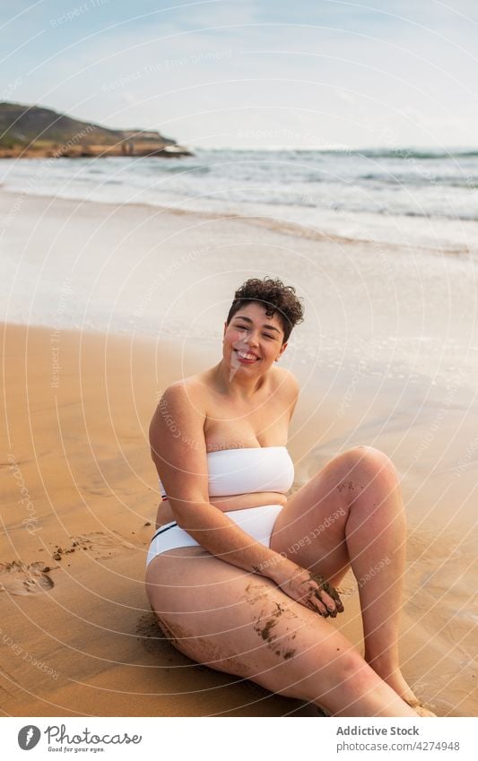 Lady sitting on sandy coast near sea woman beach ocean smile plus size swimsuit vacation blue sky happy female young aqua enjoy alone bright toothy smile