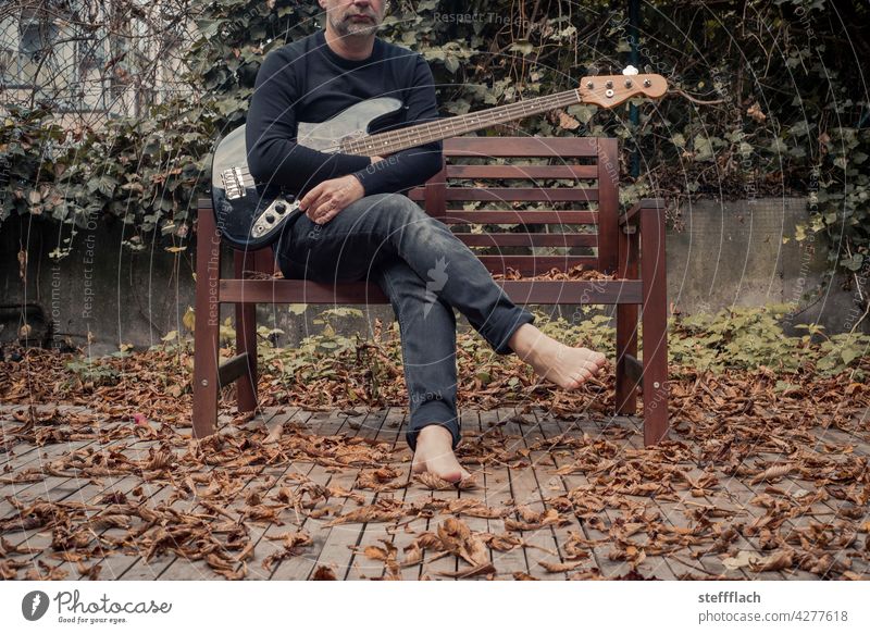 Man with electric bass and bare feet on wooden bench on terrace in autumn Autumn Terrace leaves Men in black naked feet Feet up Shriveled dried leaf