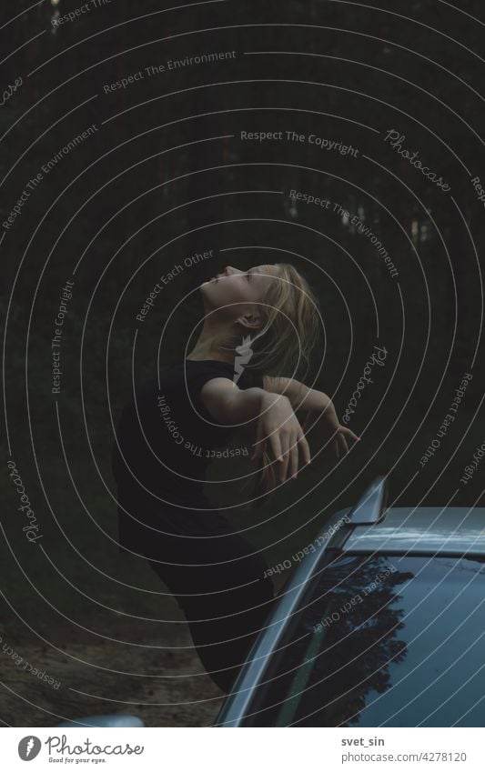 A blonde girl in black clothes keeps balance, leaning out of the window of the car, in an elegant pose with outstretched arms and thrown back head against the background of a summer twilight forest.