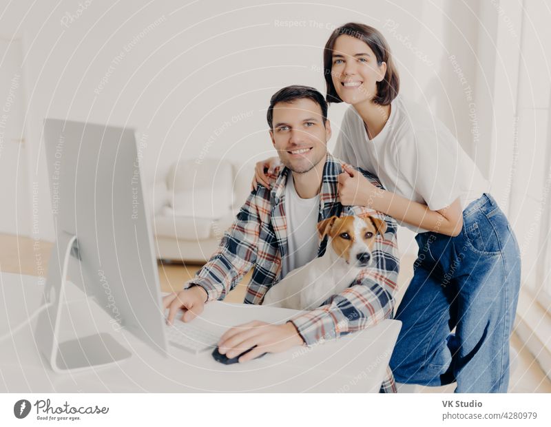 Image of happy brunette smiling woman leans at husbands shoulders who keyboards on computer, surf internet and search new apartment to buy on website, pose in spacious room with domestic dog