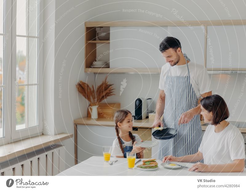 Father prepared fried eggs for family, little child holds plate and waits for breakfast. Family pose at kitchen near table, enjoy tasty meal, have glad expressions. People, eating, domestic atmosphere