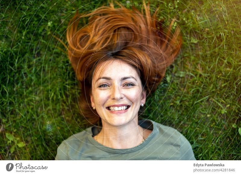 Carefree woman lying on the grass in the sun high angle daydreaming above rest green lawn meadow park nature spring red hair redhead sunny outside pretty girl