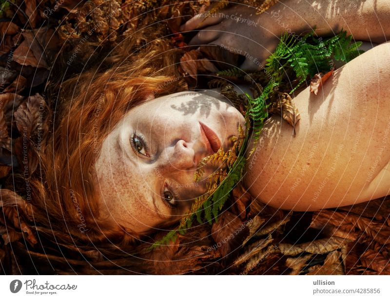 Beautiful sexy lovely young redhead girl lying on red golden autumn leaves, with a frond of fern on his chin, in the forest, with friendly seductive smile in the face