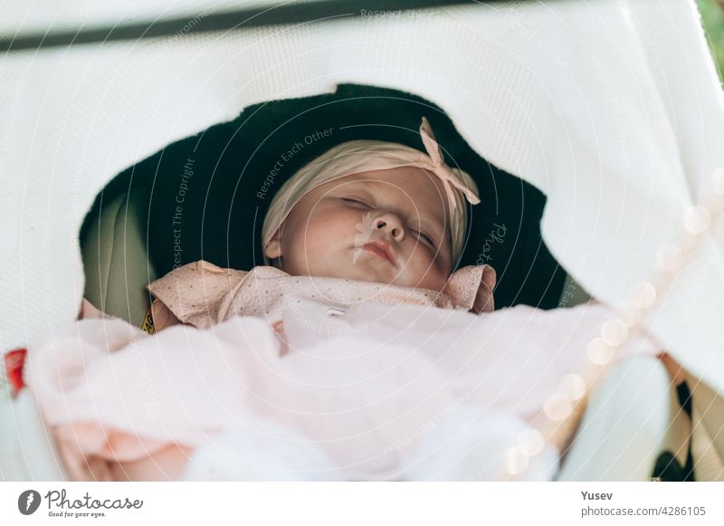 A little cute baby girl in a pink dress sleeps sweetly in stroller. Close-up. Soft focus. A beautiful infant is resting while walking in the park.Happy childhood concept. Life style photography.