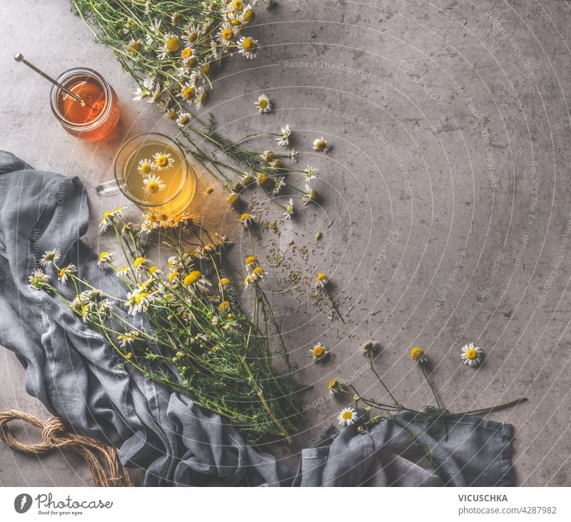 Fresh chamomile tea in glass cup, honey and honey spoon, medicinal plants. Natural treatment with herbs. Dark cloth and cord on dark concrete background. Top view.