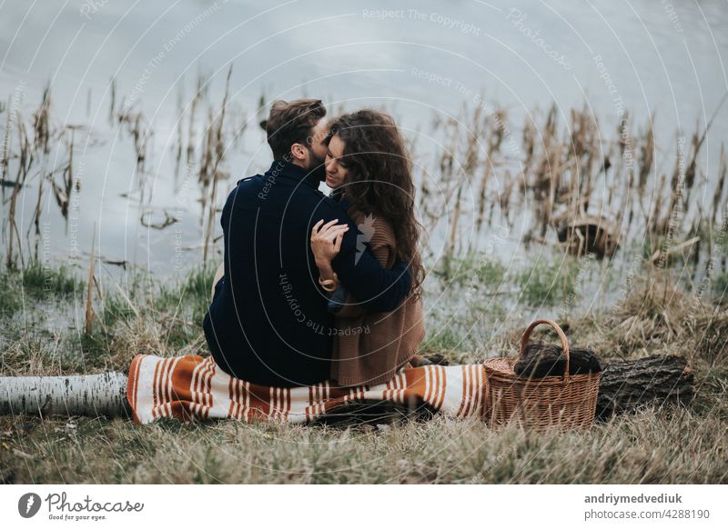 two caucasian lovers sitting on blanket by the lake. Young couple is hugging on autumn day outdoors. A bearded man and curly woman in love. Valentine's Day. Concept of love and family. back view