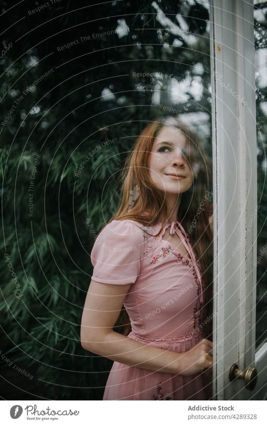 Smiling woman standing near glass doors of greenhouse garden cultivate positive charming foliage plant smile female optimist entrance vegetate growth