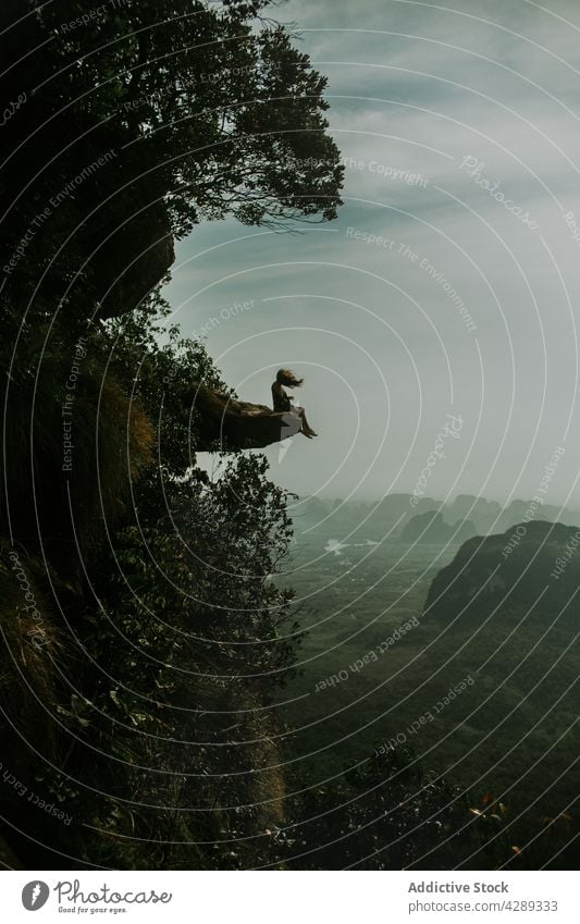 Unrecognizable woman sitting on cliff above forest backpacker rock edge jungle tree travel nature journey explore traveler green stone risk hiker rocky