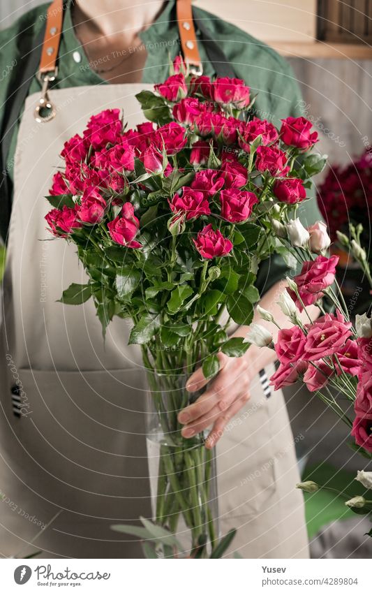 A florist woman in an apron holds a vase of roses. People at work. Florist workplace. Flower shop. Small business concept. Close-up. Vertical shot beautiful