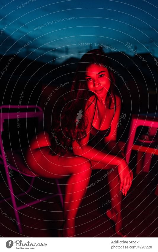 Sensual woman in lingerie sitting on terrace under neon illumination rest sensual night red light illuminate underwear chair female young balcony dark evening