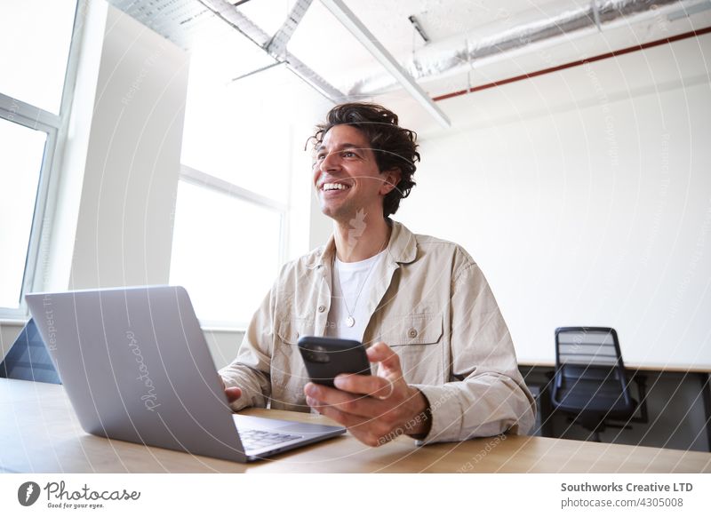 Young man with laptop and mobile phone at desk working on start-up company in open plan office Man youthful Office Desk Telephone Mobile cell review