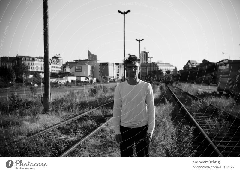 Man standing somewhat lost between disused tracks in front of city backdrop black-white Railroad tracks FALLOW LAND silent overgrown plants lost places Doomed