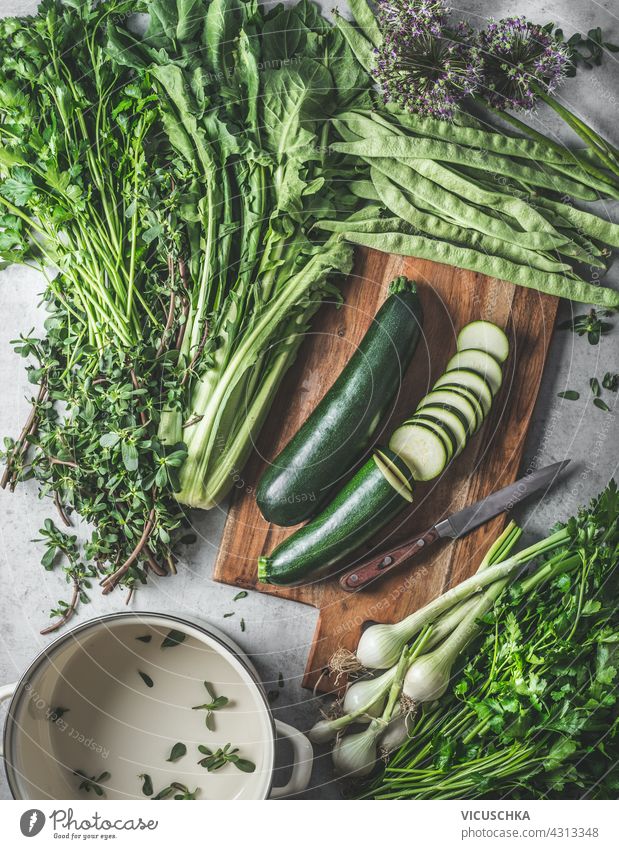 Green healthy vegetables and herbs ingredients for tasty vegan cooking on cutting board and rustic kitchen table with empty cooking pot. Top view green top view