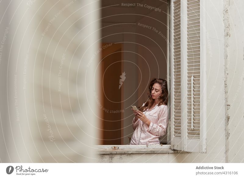 Calm woman browsing smartphone near window watching pajama conversation morning mobile female phone call cellphone device gadget nightwear sleepwear calm serene