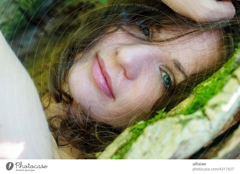 Portrait of a beautiful dark-haired young sexy woman lying happily contentedly in bed of moss in the forest with in an old willow tree with naked shoulders and deep peace in the smile