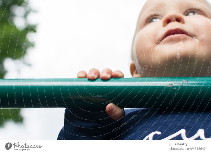 Teething toddler at the playground; baby pulling up to stand using green metal pool on equiptment teething drooling pull up balance hold grasp one year old