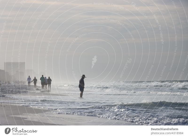 Man with hat looks at waves on the beach. Several people walking in the background. Human being person Hat short pants Jacket Beach Sand Waves in the morning