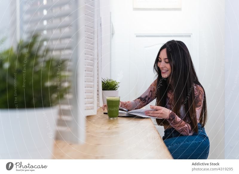 Delighted woman taking notes in notebook in bar with cocktail drink take note write beverage enjoy chill female smile counter cheerful happy sit glass notepad