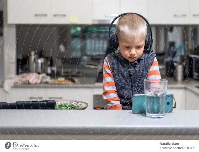 Little child boy wares headphone and playing the cell phone on the kitchen table computer son tablet mobile phone studying cook house internet technology