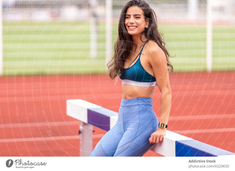 Happy sportswoman resting near hurdle on stadium track athlete happy smile break sportswear runner female young hispanic ethnic training workout fitness healthy