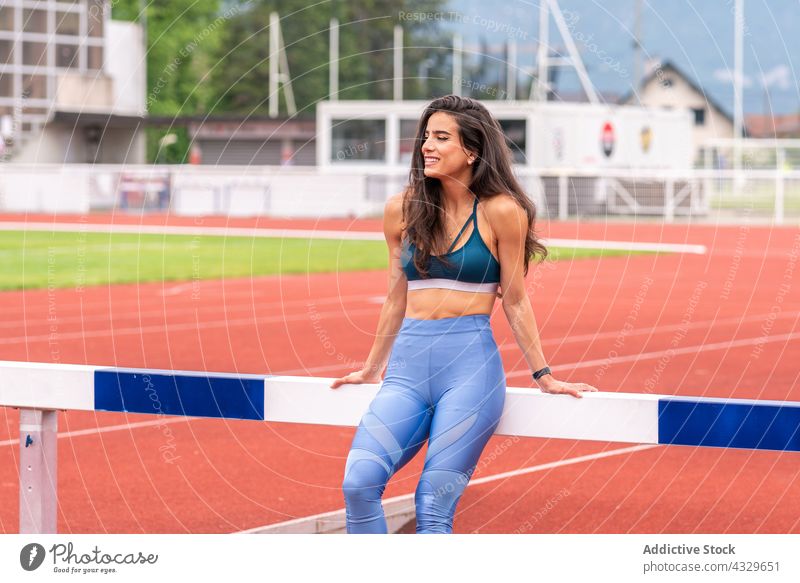 Happy sportswoman resting near hurdle on stadium track athlete happy smile break sportswear runner female young hispanic ethnic training workout fitness healthy