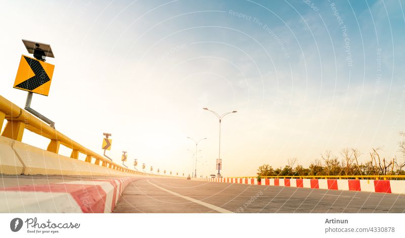 Curve concrete road with curve traffic sign beside the sea at sunset time. Solar panel energy on yellow curve traffic sign. Road trip on summer vacation. Blur car driving. Summer travel by car.