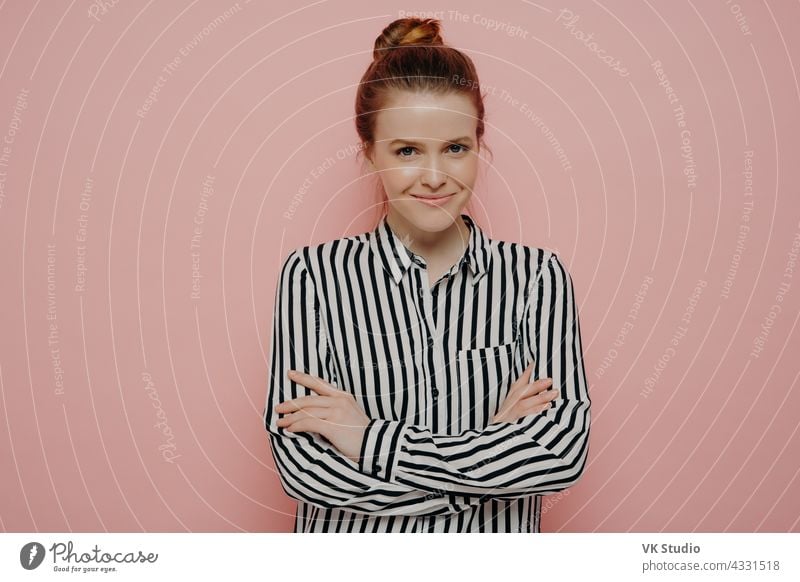 Happy teenage girl with crossed arms posing against pink studio background satisfied happy redhead woman hair bun looking straight smiling joyfully camera