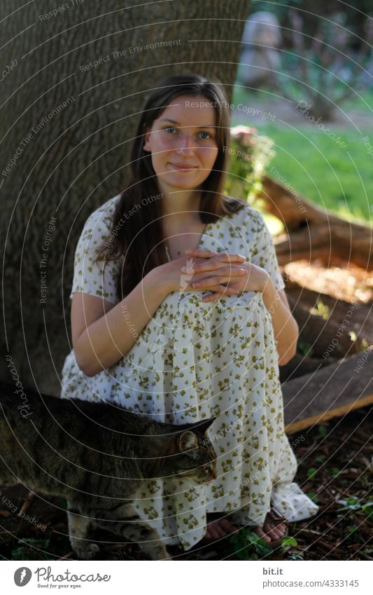 Young natural, long-haired woman sits on holiday, in summer dress, with her tiger cat in the garden, in her favorite place, by the tree in the shade.