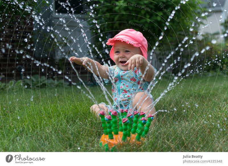 One year old baby happily playing in sprinkler; simple water play entertainment for toddler sun safety yard home neighborhood midwest spf upf sun block clothing