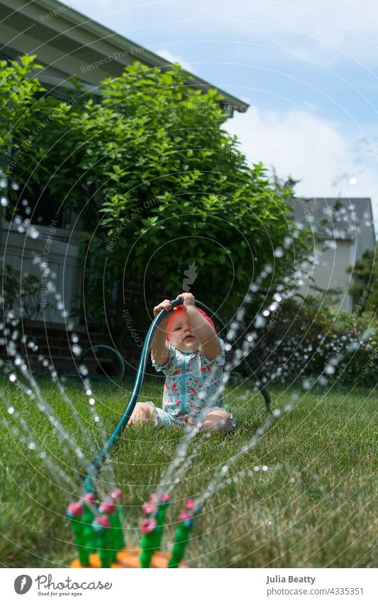 Young toddler hold onto garden hose while playing in sprinkler in front yard baby sun safety fine motor pull water water play home neighborhood midwest spf upf