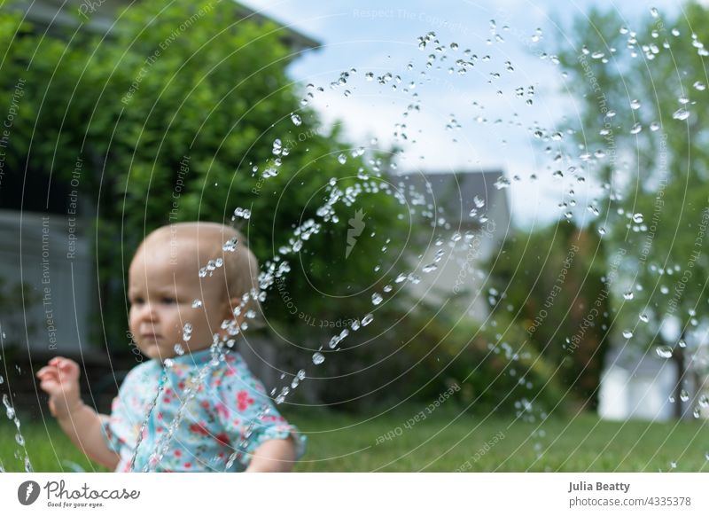 Toddler seated in grass in front yard; in front of her a sprinkler sprays water baby toddler sun safety water play home neighborhood midwest spf upf