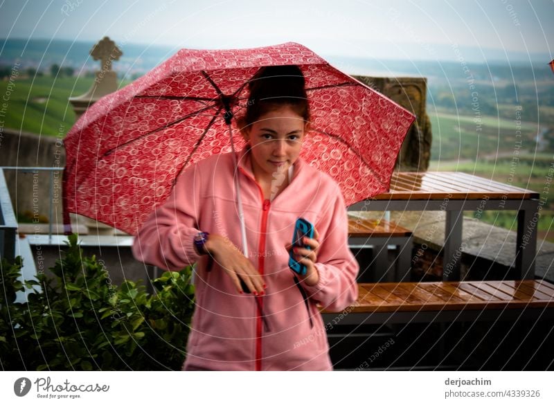 Everything in pink. Young girl stands with umbrella and rain jacket in the outdoor area of the restaurant.  The mobile phone in hand and looks desperately to the photographer, when is he finally ready .