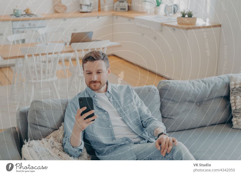 Attractive caucasian young man in domestic casual clothes using smartphone at home bearded cheerful mobile smiling sofa person positivity rest resting surfing